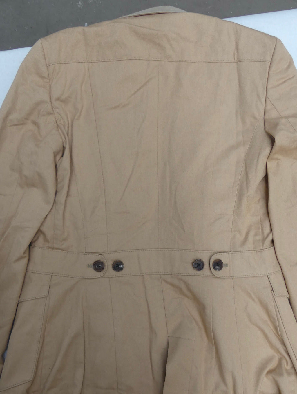A tan utility jacket shown from the back, featuring a structured collar, long sleeves, and a waist belt with dark buttons.