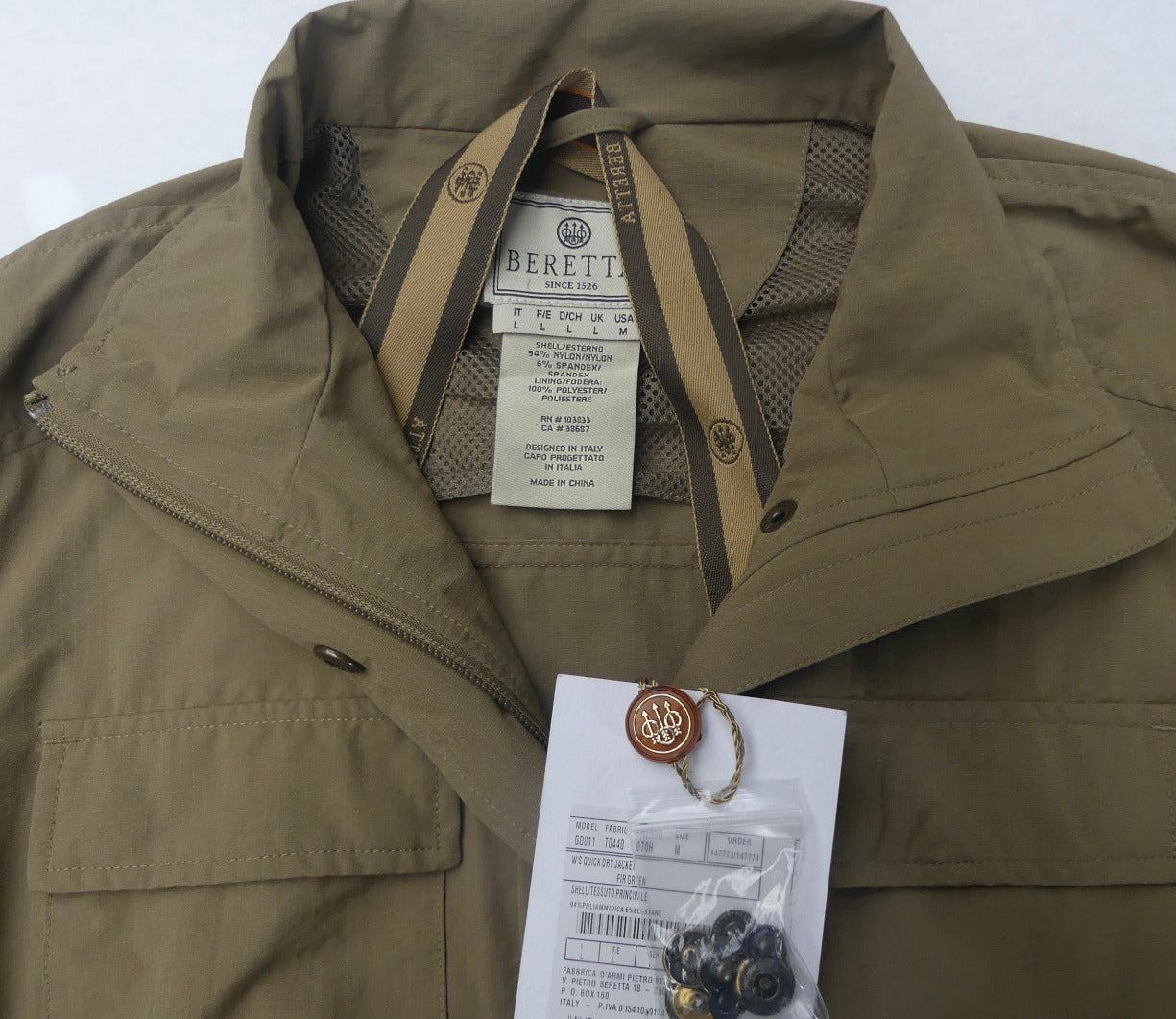 A close-up of a Beretta jacket in olive drab green, showing the collar, zipper, interior lining, and attached hangtag with a small accessory inside.