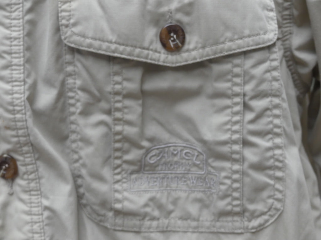 Close-up of a light beige or off-white button-up shirt or jacket pocket with a visible embroidered 'Camel' logo patch on the front flap.