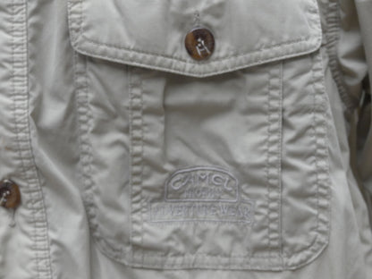 Close-up of a light beige or off-white button-up shirt or jacket pocket with a visible embroidered 'Camel' logo patch on the front flap.