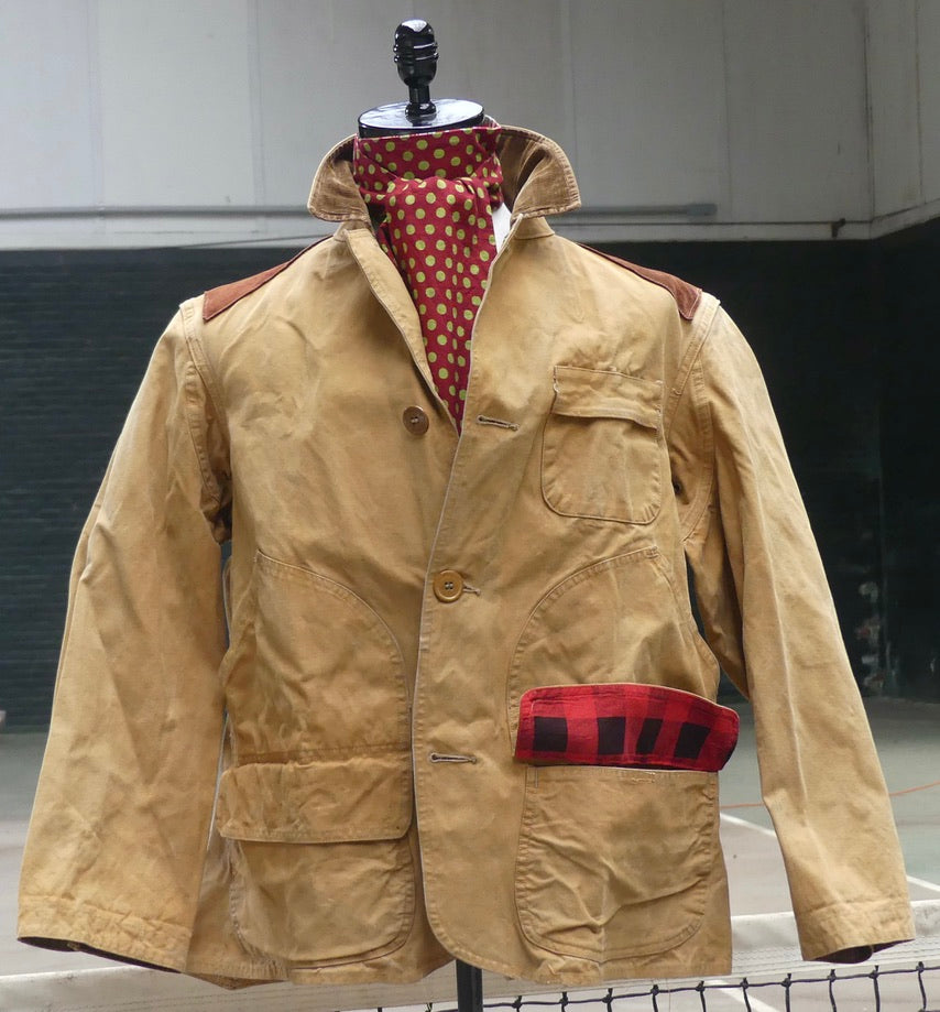 A tan waxed canvas field jacket displayed on a mannequin, featuring brown leather shoulder patches, a red and white polka dot neck scarf, and a red and black buffalo plaid pocket flap.