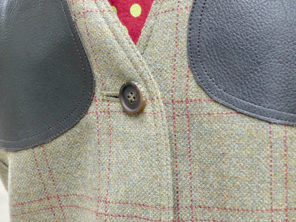 Close-up of a tweed jacket with dark gray leather wing patches and a brown button, featuring a beige and gray check pattern with red windowpane lines.