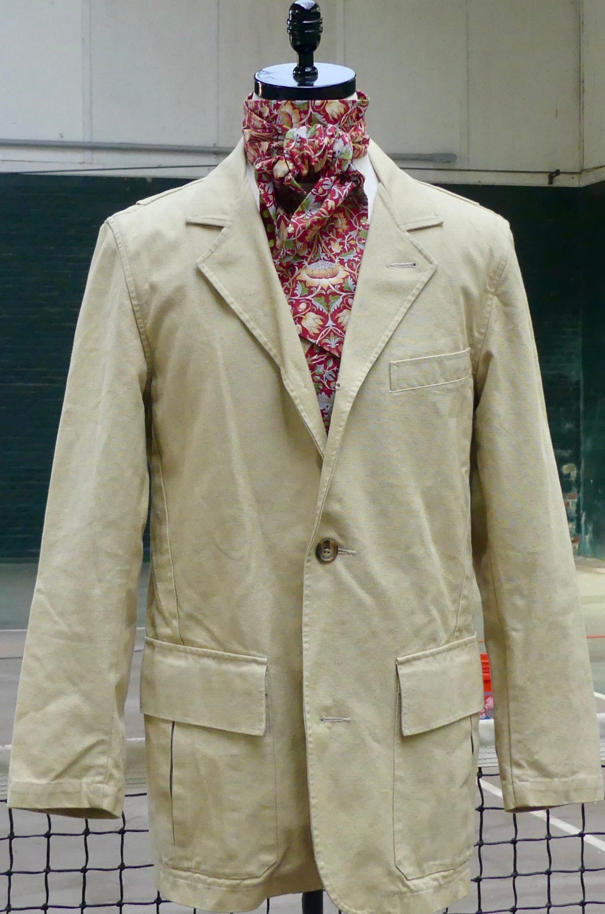 A beige men's blazer jacket displayed on a mannequin torso, styled with a red and gold patterned neck scarf. The jacket features a single button closure, notch lapels, and two large front flap pockets.
