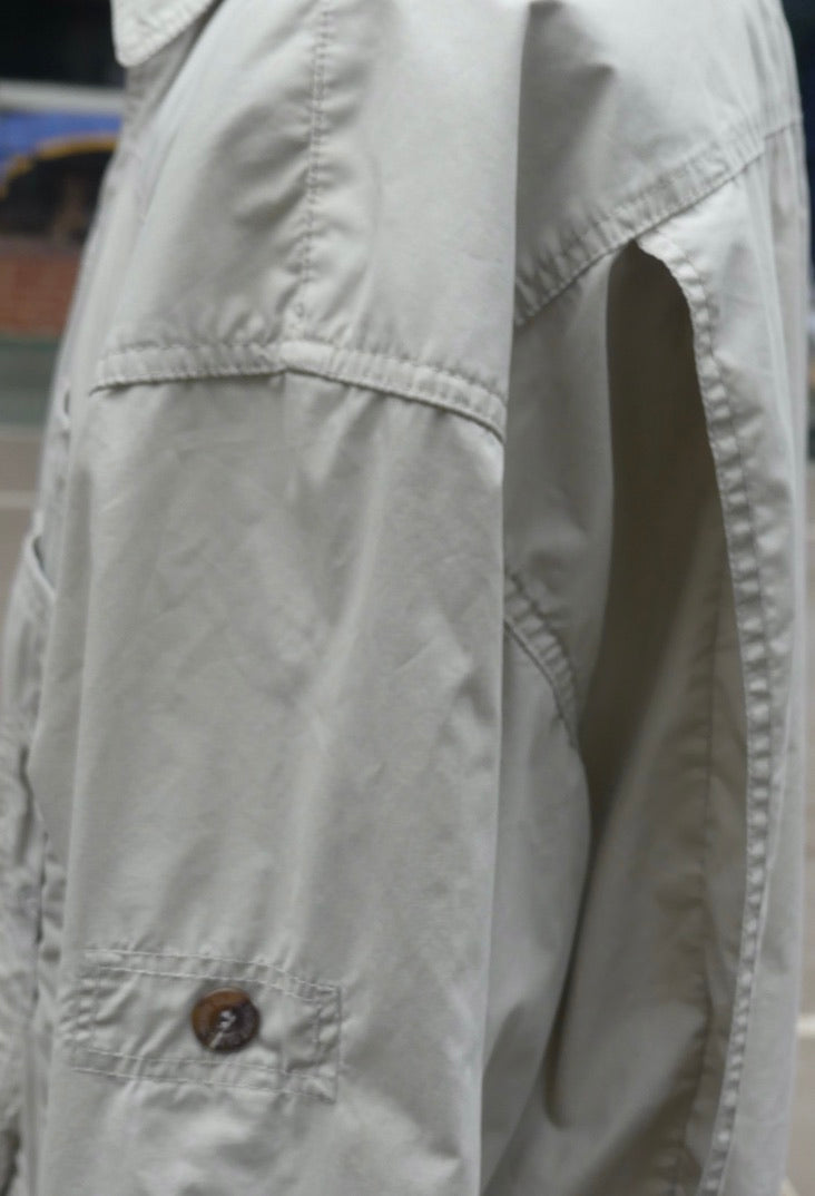 Close-up of a light beige or off-white utility jacket with a brown button on a pocket, showing detailed fabric stitching and texture.