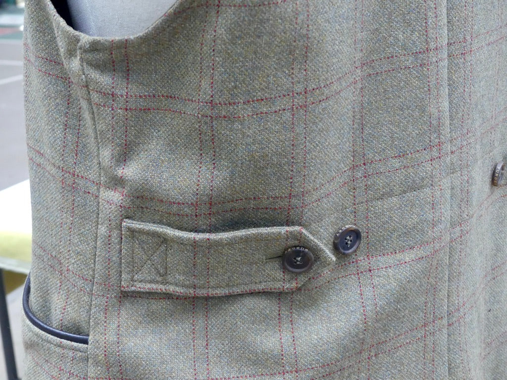 Close-up of a tweed waistcoat in a light gray and beige check pattern with thin red lines, featuring a buttoned flap pocket and dark buttons.
