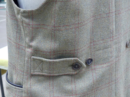 Close-up of a tweed waistcoat in a light gray and beige check pattern with thin red lines, featuring a buttoned flap pocket and dark buttons.