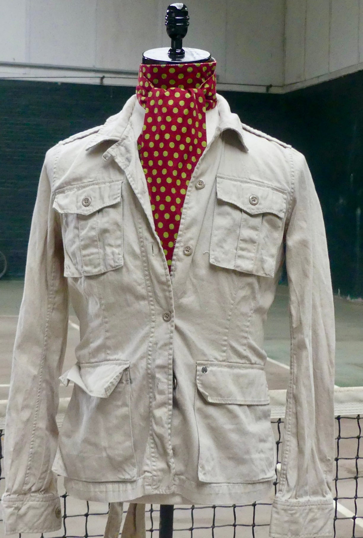 A light beige military-style jacket displayed on a mannequin torso, styled with a red neck scarf featuring a gold polka dot pattern. The jacket has four front flap pockets and is shown against an industrial background.