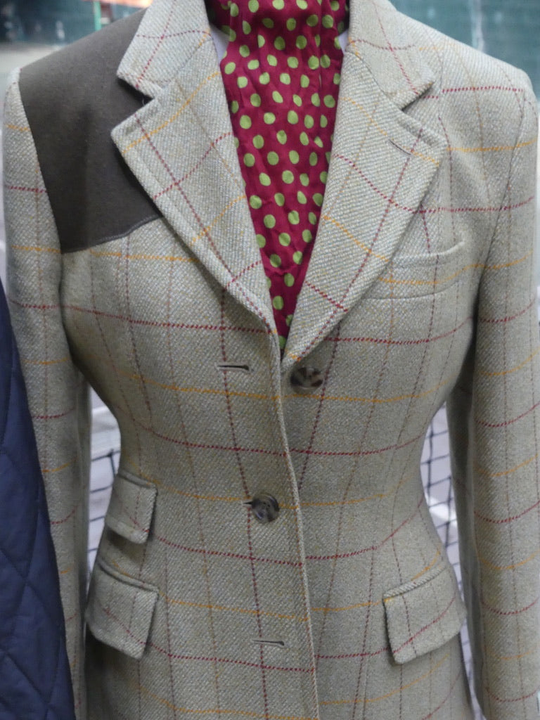 A tailored tweed blazer in a light beige-gray with a red and yellow windowpane plaid pattern, featuring dark gray shoulder panels and a red polka-dot turtleneck underneath.