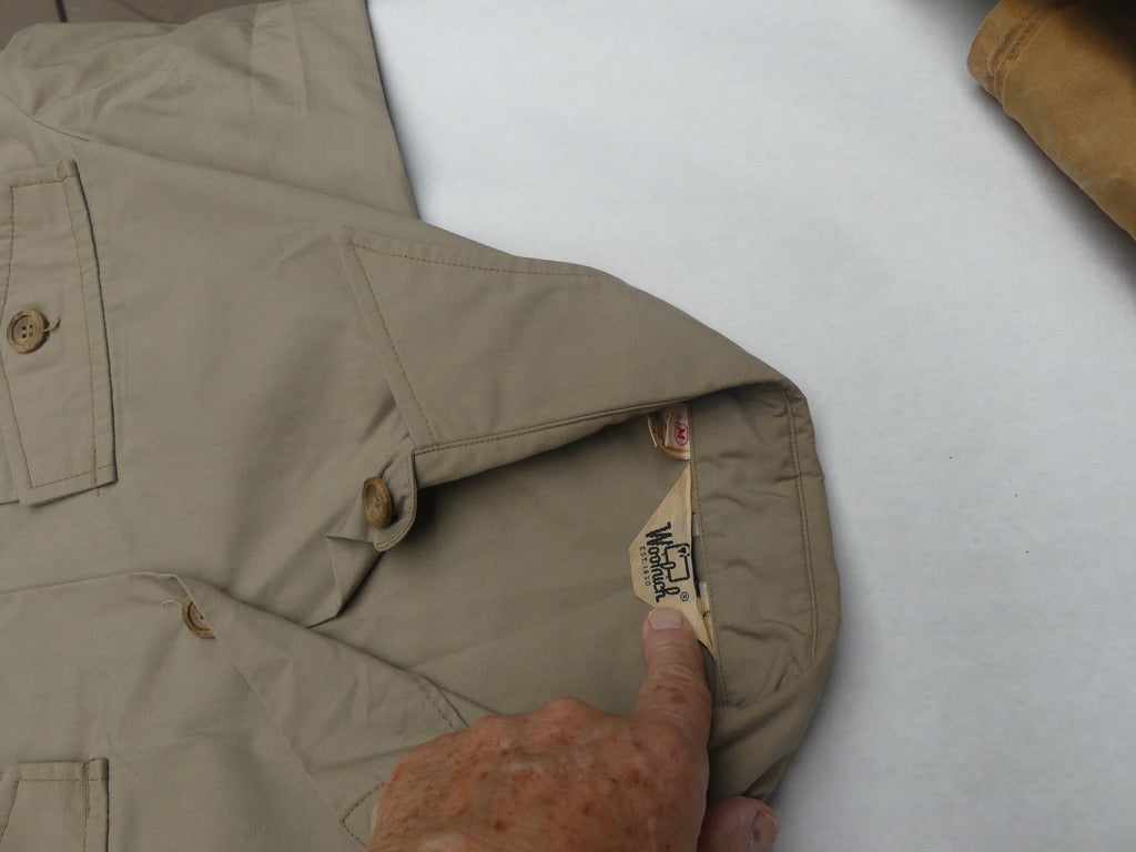 A close-up of a khaki beige utility shirt laid flat on a white surface, with a hand pointing to the inner neck label which reads 'Mackay'.