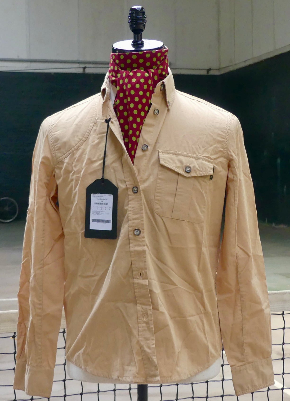 A beige long-sleeve button-up shirt displayed on a mannequin torso, styled with a red and gold polka dot neck scarf. The shirt features a chest pocket with a snap button and a black hangtag attached.