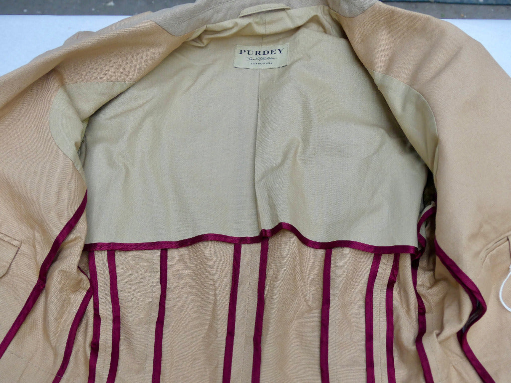 A close-up of a beige and tan jacket or coat laid flat, showing the interior lining and a magenta-pink striped lining detail along the lower front. A neck label reads PURDEY.