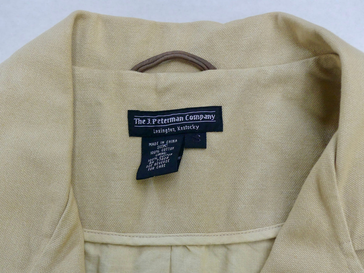Close-up of the inside collar of a light beige woven garment, showing a black fabric label that reads 'The J Peterman Company' with 'Lexington, Kentucky' underneath.